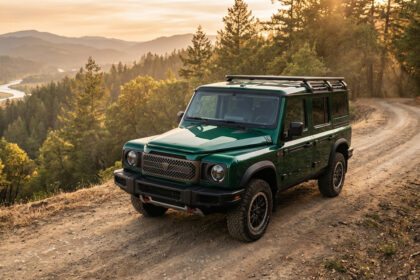 SUV Ineos Grenadier Chelsea vert foncé sur chemin terre. Vallée boisée, rivière au loin, lumière dorée du coucher du soleil.