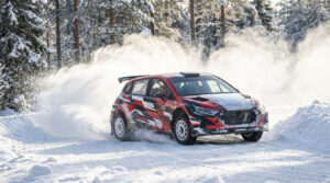 Voiture de rallye rouge, noire et blanche glissant sur une piste enneigée, soulevant de la neige dans une forêt hivernale.