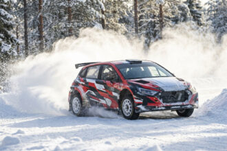 Voiture de rallye rouge, noire et blanche glissant sur une piste enneigée, soulevant de la neige dans une forêt hivernale.