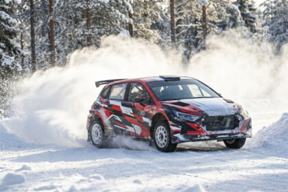 Voiture de rallye rouge, noire et blanche glissant sur une piste enneigée, soulevant de la neige dans une forêt hivernale.