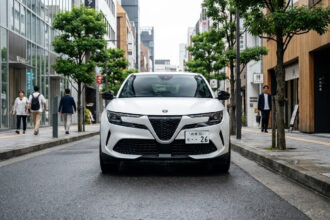 Alfa Romeo Junior blanc garé au centre d'une rue urbaine japonaise mouillée, entouré de piétons et de bâtiments modernes.