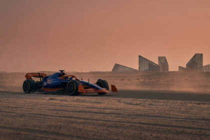 Voiture de Formule 1 bleue et orange sur une piste désertique sous un ciel ocre, avec des bâtiments modernes en arrière-plan.