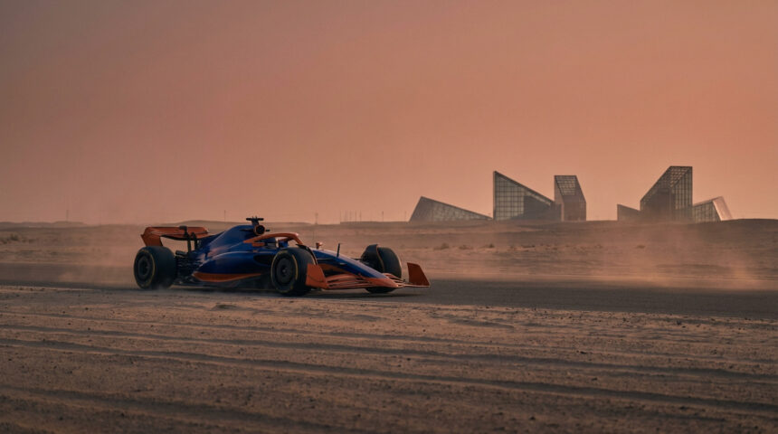 Voiture de Formule 1 bleue et orange sur une piste désertique sous un ciel ocre, avec des bâtiments modernes en arrière-plan.