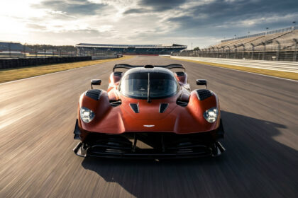 Aston Martin Valkyrie AMR Pro orange foncée sur une piste de course, effet de vitesse. Tribunes vides à l'arrière-plan sous un ciel nuageux.