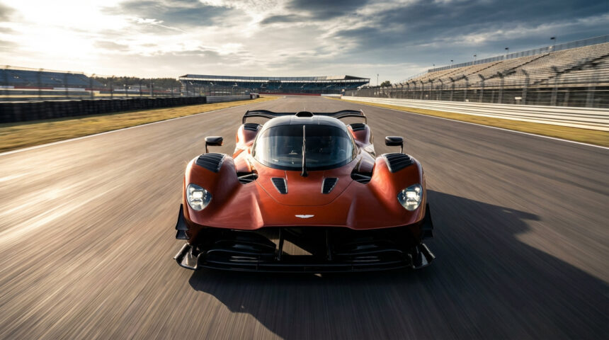 Aston Martin Valkyrie AMR Pro orange foncée sur une piste de course, effet de vitesse. Tribunes vides à l'arrière-plan sous un ciel nuageux.