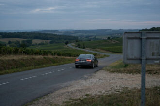 Voiture sombre sur route sinueuse traversant des vignobles sous un ciel nuageux au crépuscule, panneau de dos à droite.