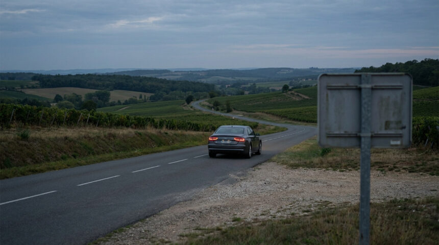Voiture sombre sur route sinueuse traversant des vignobles sous un ciel nuageux au crépuscule, panneau de dos à droite.