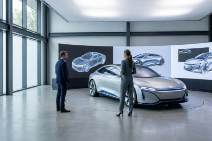 Deux personnes face à une voiture concept argentée dans un studio. Des écrans affichent des dessins et modèles 3D du véhicule.