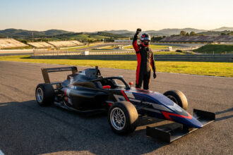 Un pilote de course en tenue et casque lève le poing en signe de victoire à côté d'une monoplace sur un circuit.