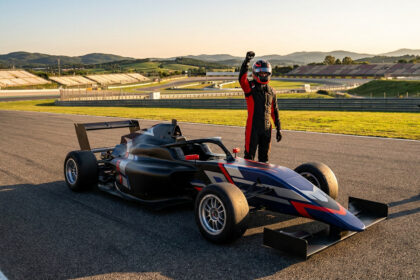Un pilote de course en tenue et casque lève le poing en signe de victoire à côté d'une monoplace sur un circuit.