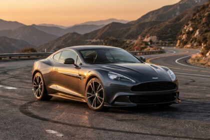 Une Aston Martin Vanquish 25 grise garée sur une route sinueuse en montagne au coucher du soleil.