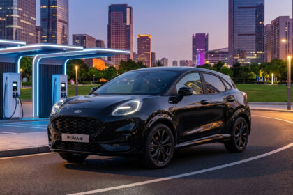 Ford Puma Gen-E noir stationné à une borne de recharge électrique devant des gratte-ciel urbains au crépuscule.