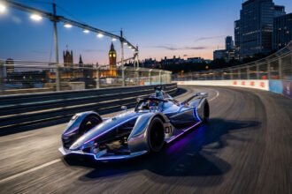 Une monoplace de Formule E chromée avec des bandes lumineuses circulant sur le circuit urbain de Londres au crépuscule.