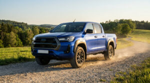 Un pick-up Toyota Hilux bleu roulant sur un chemin de terre poussiéreux dans un paysage rural ensoleillé.