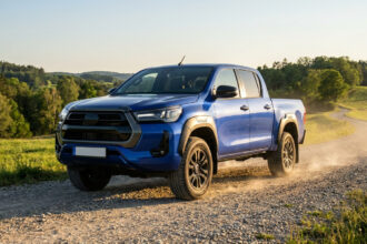 Un pick-up Toyota Hilux bleu roulant sur un chemin de terre poussiéreux dans un paysage rural ensoleillé.