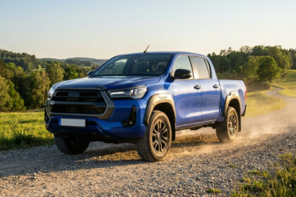 Un pick-up Toyota Hilux bleu roulant sur un chemin de terre poussiéreux dans un paysage rural ensoleillé.
