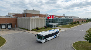 Un autobus électrique blanc circulant devant l'usine de Brampton sous le drapeau canadien.