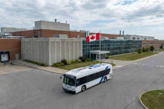 Un autobus électrique blanc circulant devant l'usine de Brampton sous le drapeau canadien.