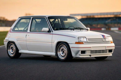 Renault 5 GT Turbo blanche de compétition avec bandes bleues et rouges sur un circuit de course au coucher du soleil.
